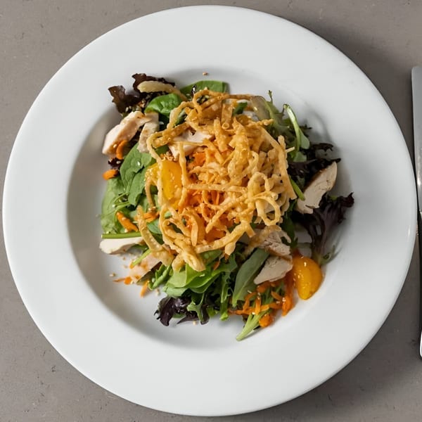 Overhead view of a vibrant chicken salad with greens, mandarins, and crispy noodles on a white plate.