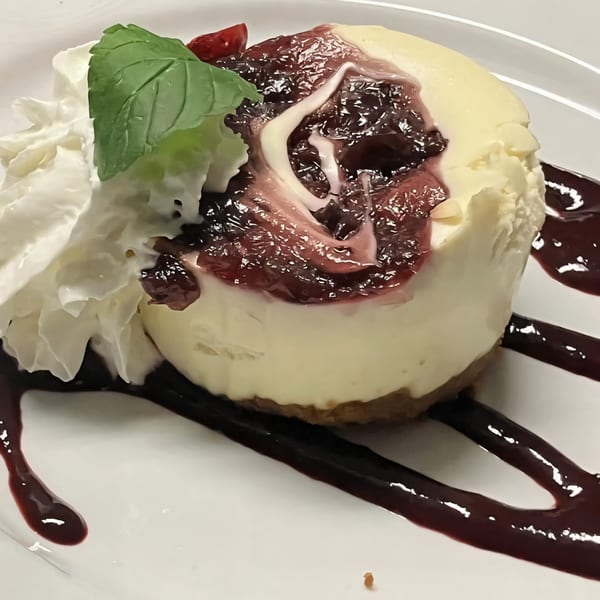 Cheesecake with berry compote, whipped cream, and a mint leaf on a white plate.