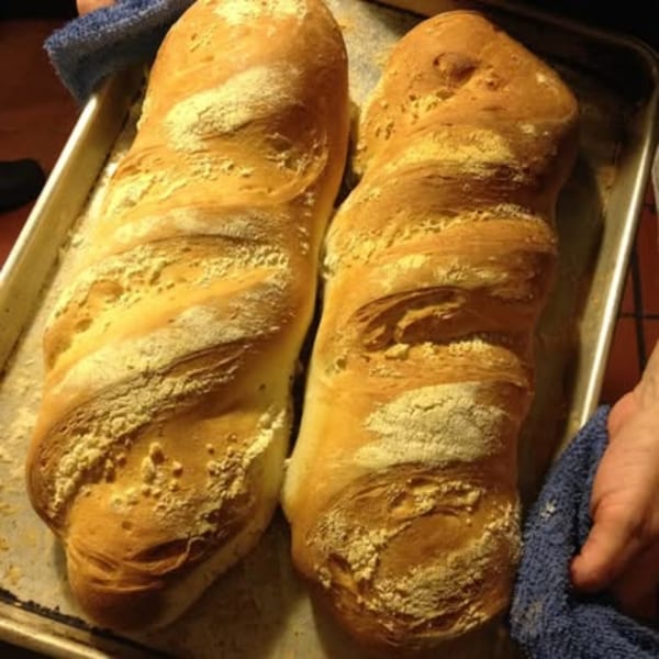 Two freshly baked loaves of golden-brown bread, dusted with flour, on a baking sheet.