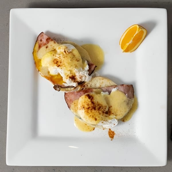 Eggs Benedict with ham, hollandaise sauce, and a side of orange on a white square plate.